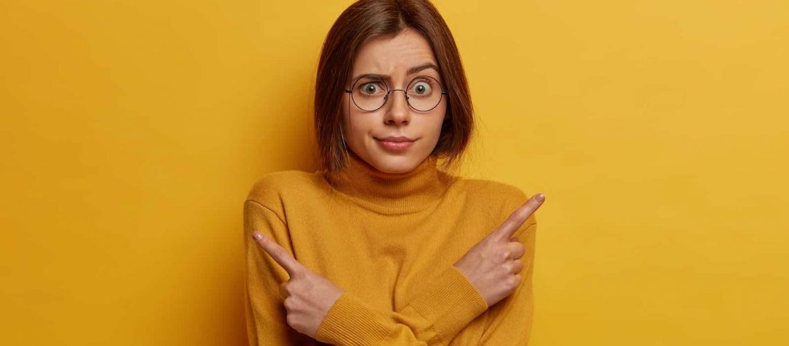 Studio shot of embarrassed unaware woman points sideways, thinks what to choose, raises eyebrows, indicates right and left, dressed in yellow outfit, stands indecisive indoor. Difficult choice