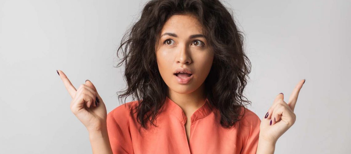 portrait of young pretty woman, mixed race, curly hair, suprised, shocked emotion on face, looking in camera, isolated, orange blouse, big eyes, open mouth, pointing fingers up, thinking about choise