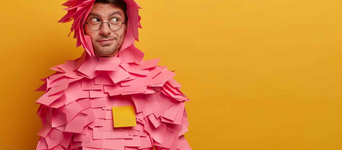 Indoor shot of male advertiser or manager covered with sticky adhesive notes, looks aside as notices something interesting, poses against yellow background, free space for your advertising content
