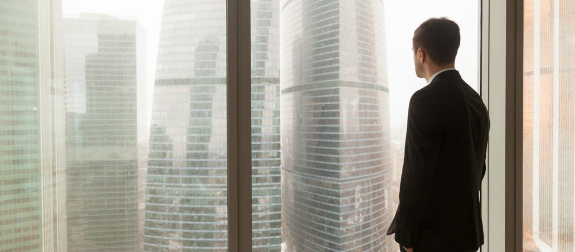 Homme en costume d'affaires debout, les mains dans les poches, regardant par la fenêtre un paysage urbain avec des gratte-ciel. Homme d'affaires pensant aux perspectives, rêvant de la réussite de l'entreprise. Vue arrière