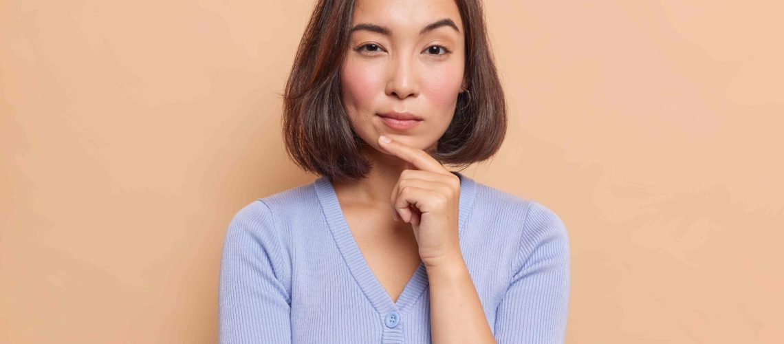 Portrait of serious dark haired Asian woman keeps finger on chin looks mysteriously at camera considers something dressed in blue jumper isolated over brown background. Let me think about it